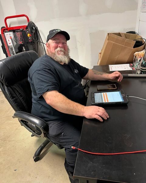 Man with beard at a desk, looking at tablet. Wearing black shirt and cap in a workshop.