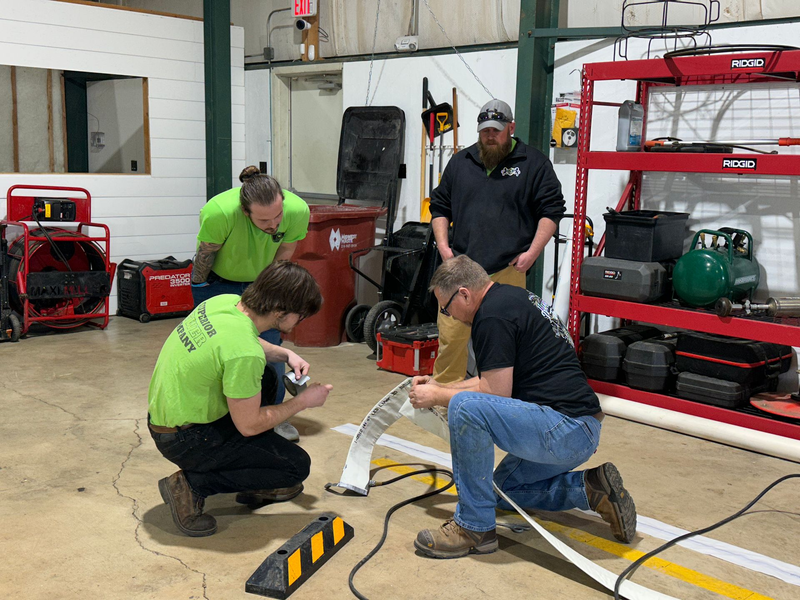 Four men in a garage working on a wire. One is kneeling, others standing and observing. Tools and equipment visible.