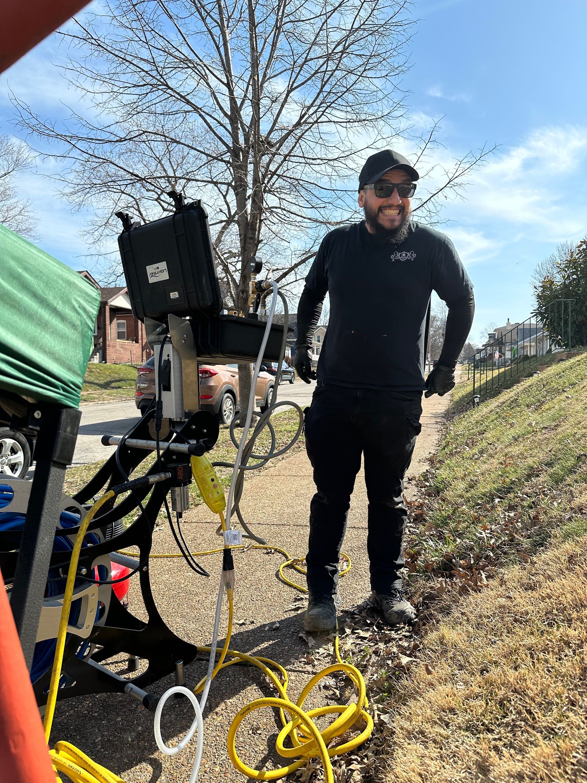Man in black clothing stands near equipment on a sunny day; possibly filming outdoors.