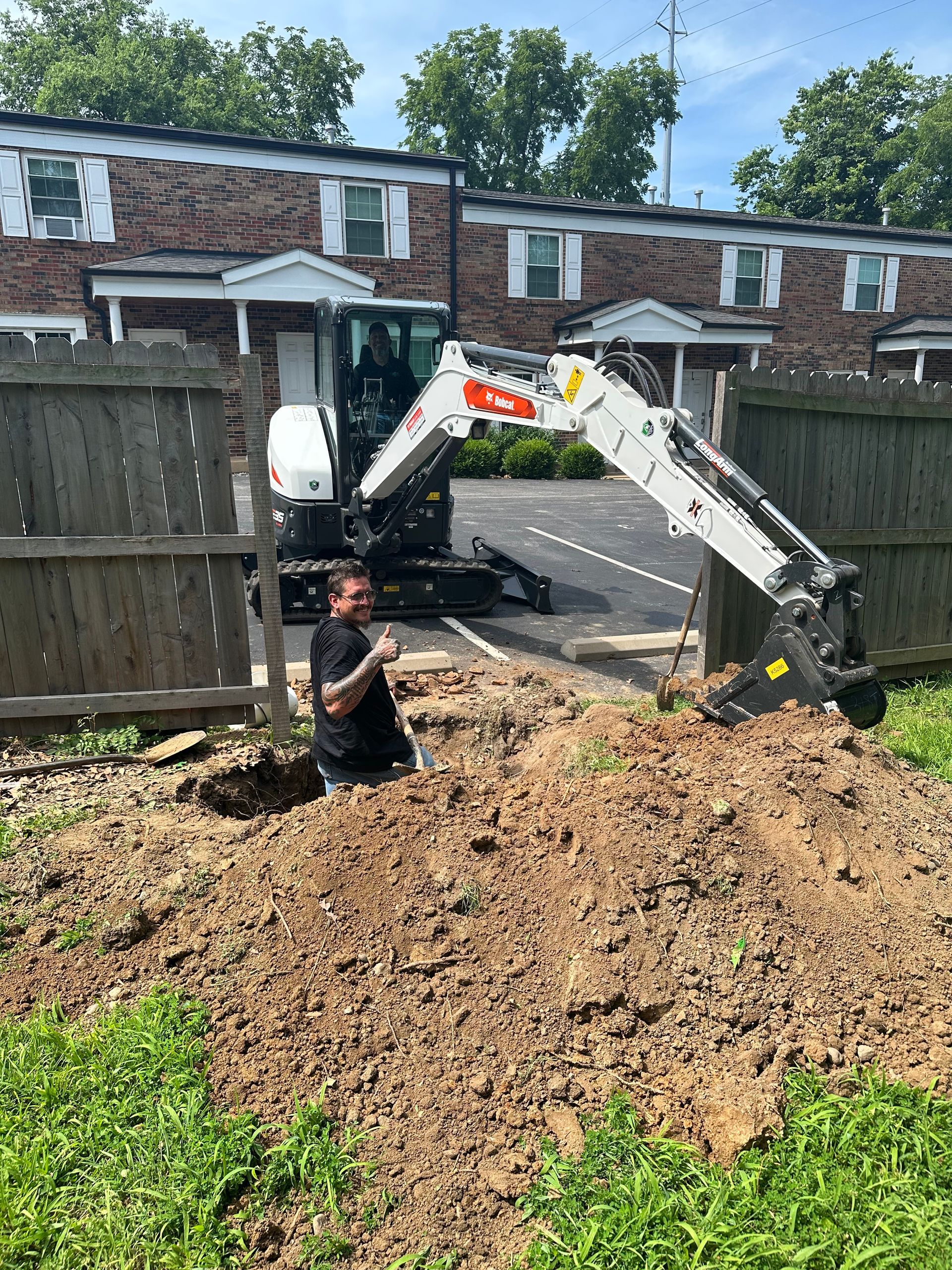 Man operating a Bobcat excavator digging near a fence in front of an apartment building.