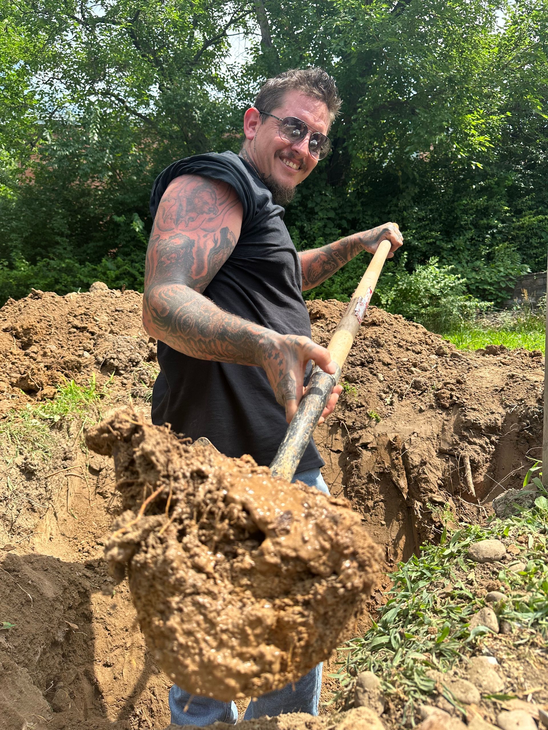 Man with tattoos smiles, holding a shovel full of dirt in a muddy, outdoor area.