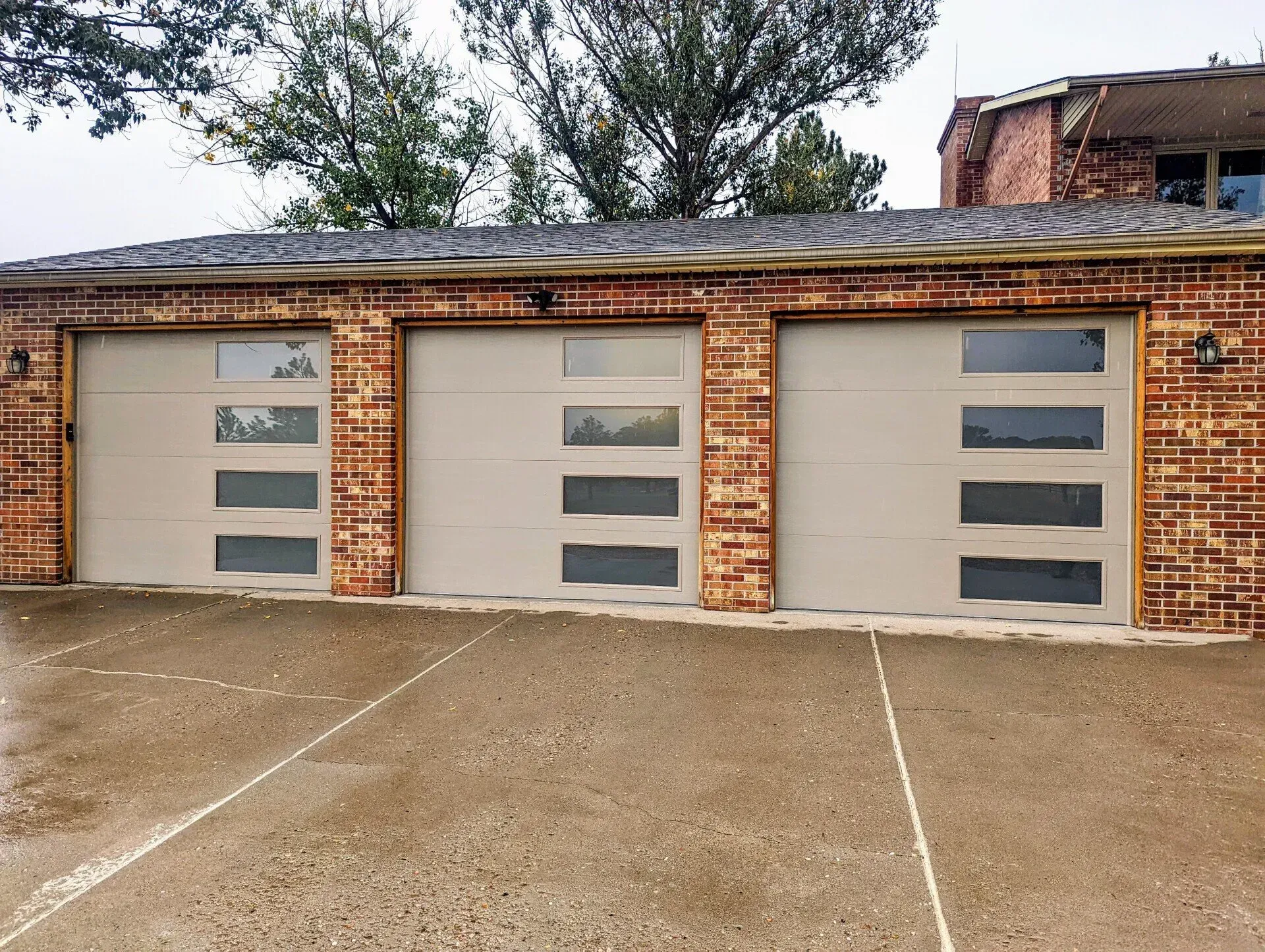 Three tan garage doors with glass panels set in a red brick building, concrete driveway.