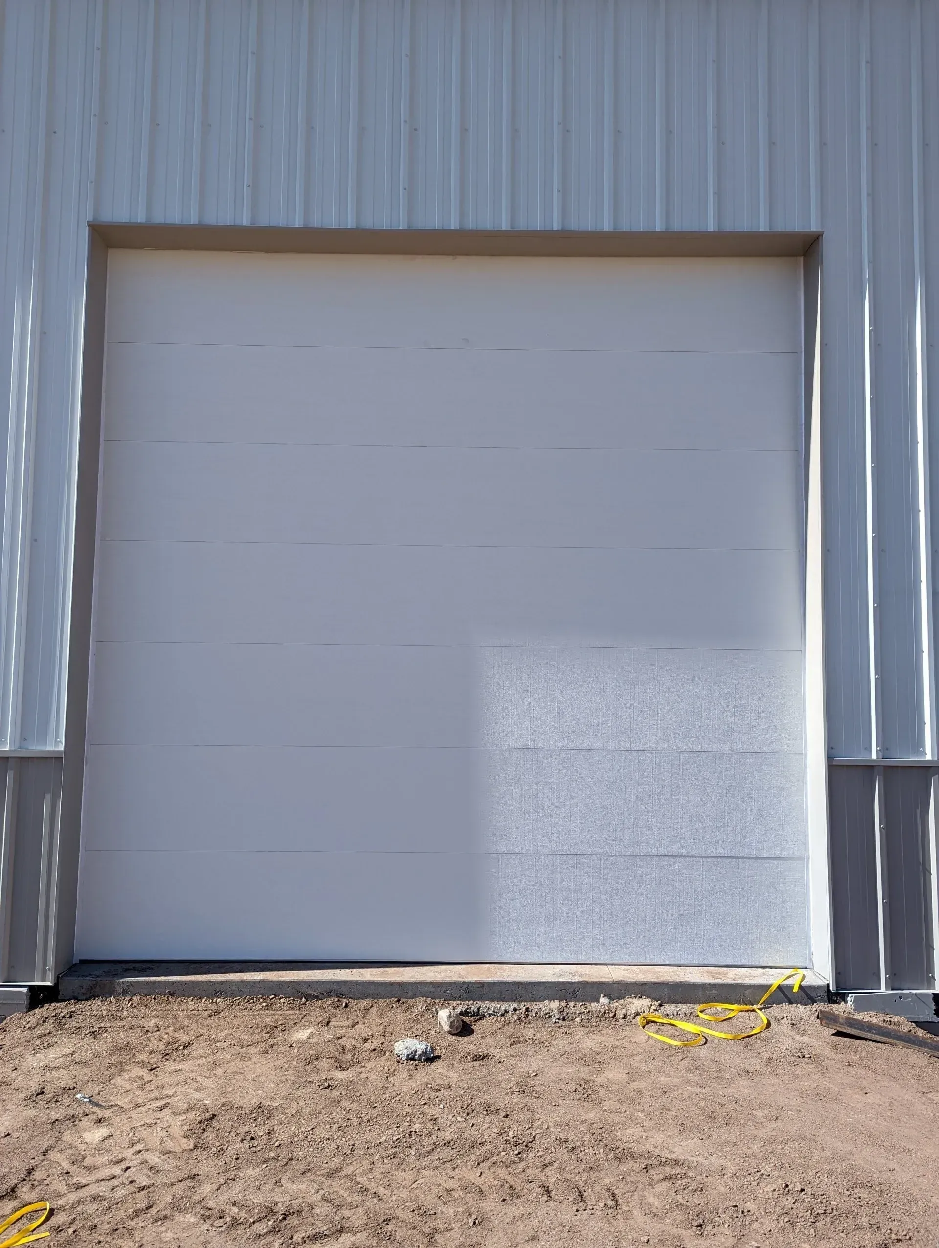White closed garage door in a light gray metal building, set in a dirt lot.