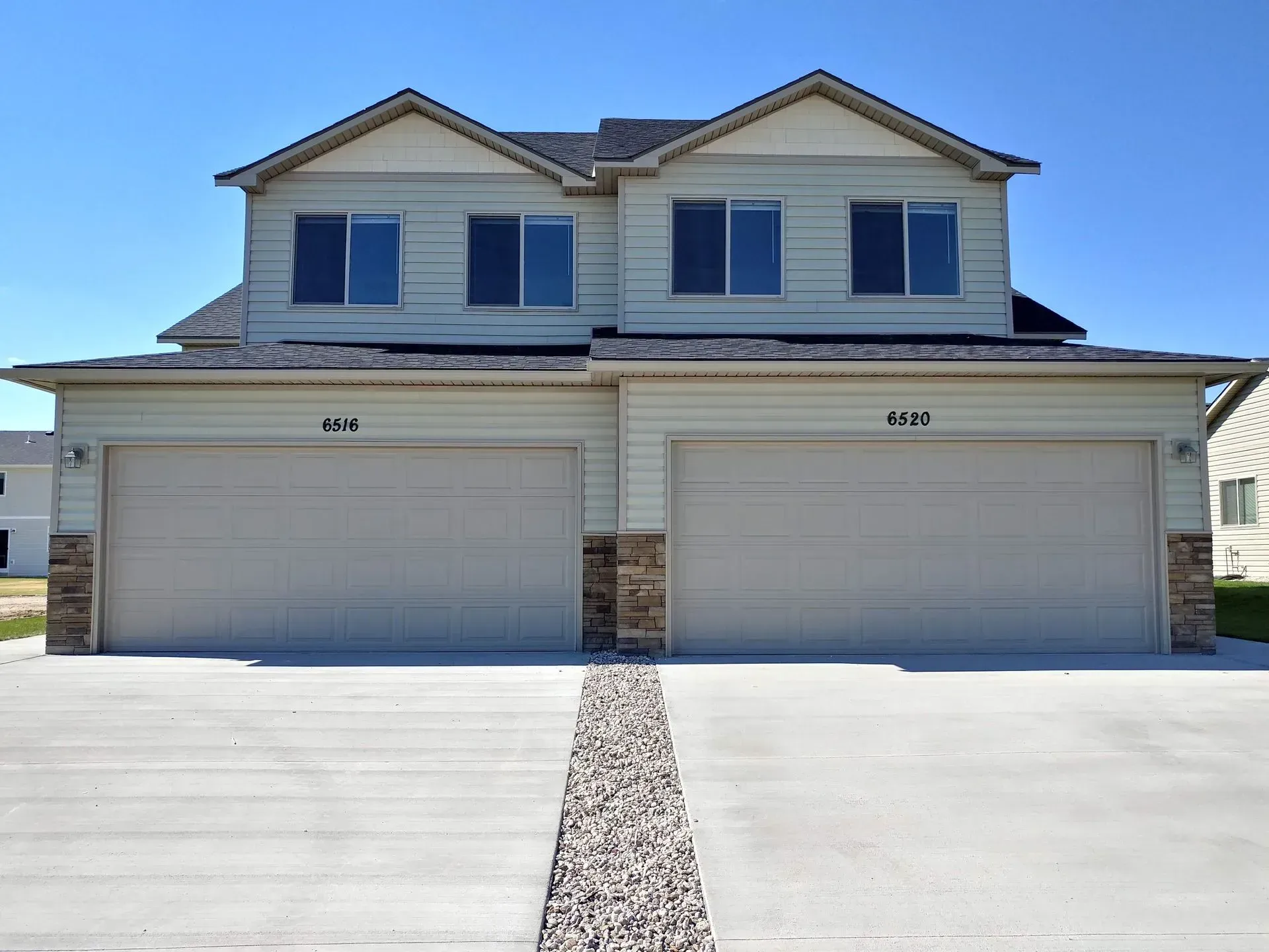 Duplex with two-car garages and light siding, on a sunny day. Driveway with stone border, blue sky.