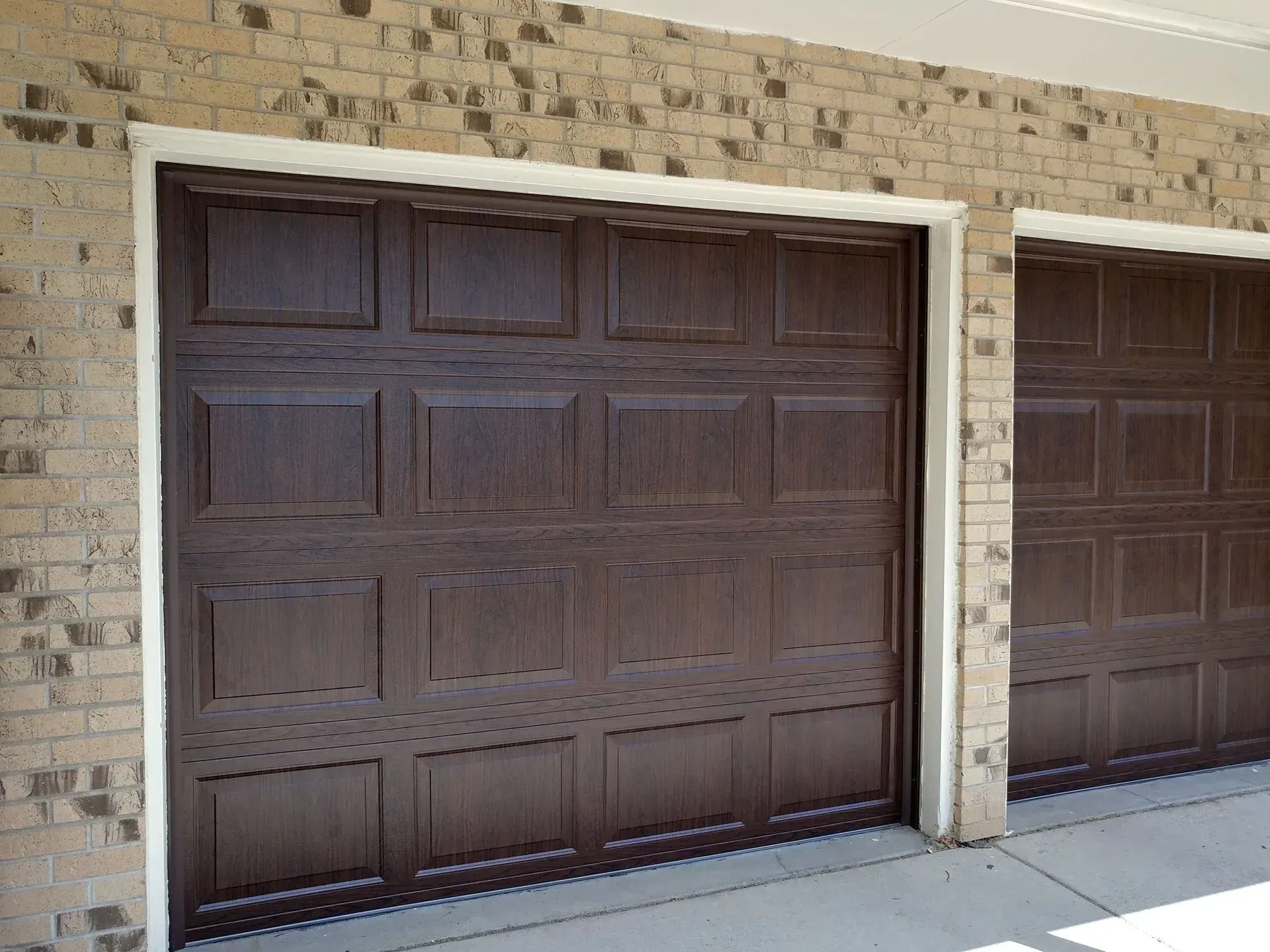 Two dark brown garage doors with panel details against a brick wall, both framed in white.