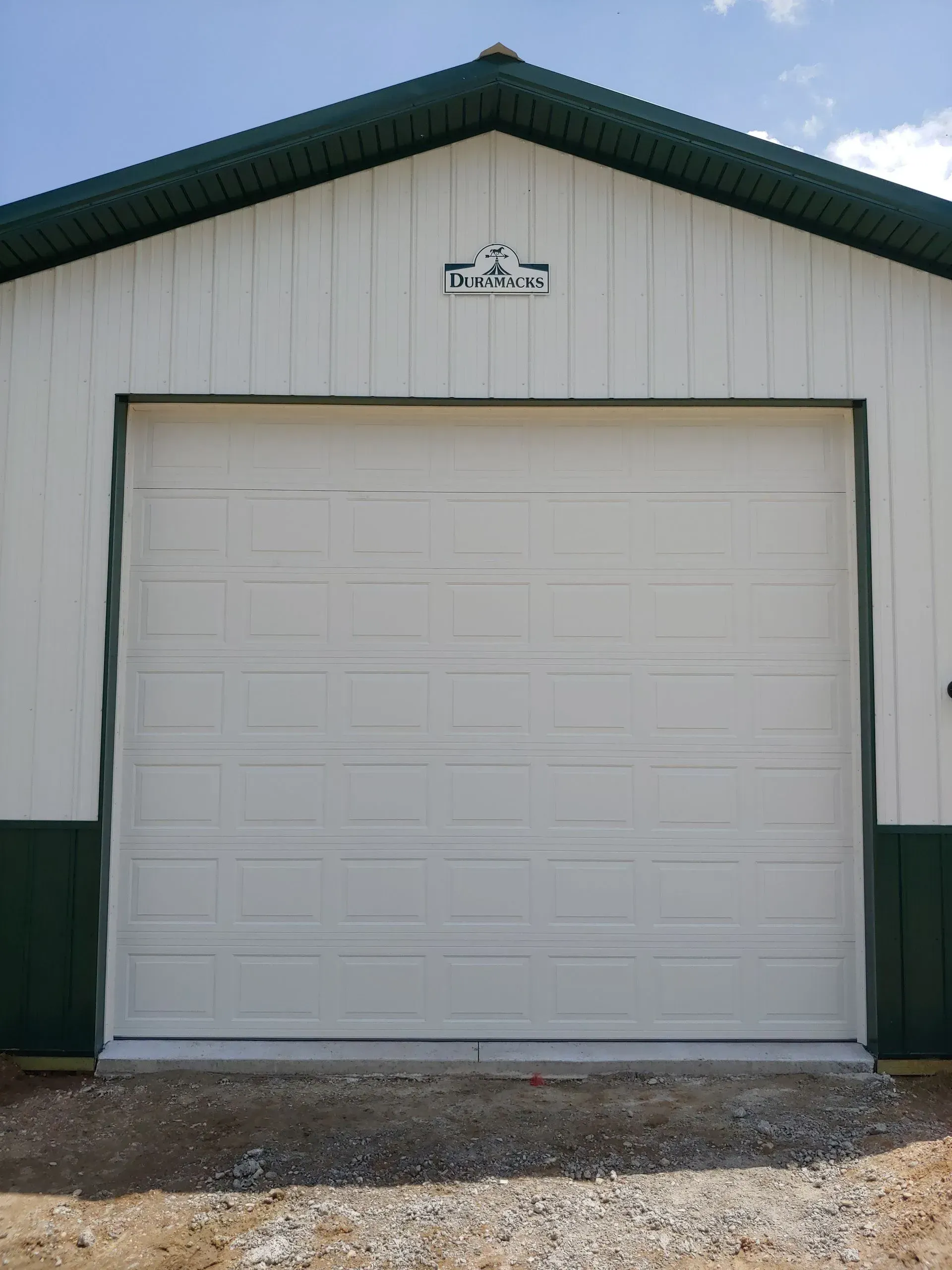 White garage door on a white and green building with a green roof, on a gravel surface.