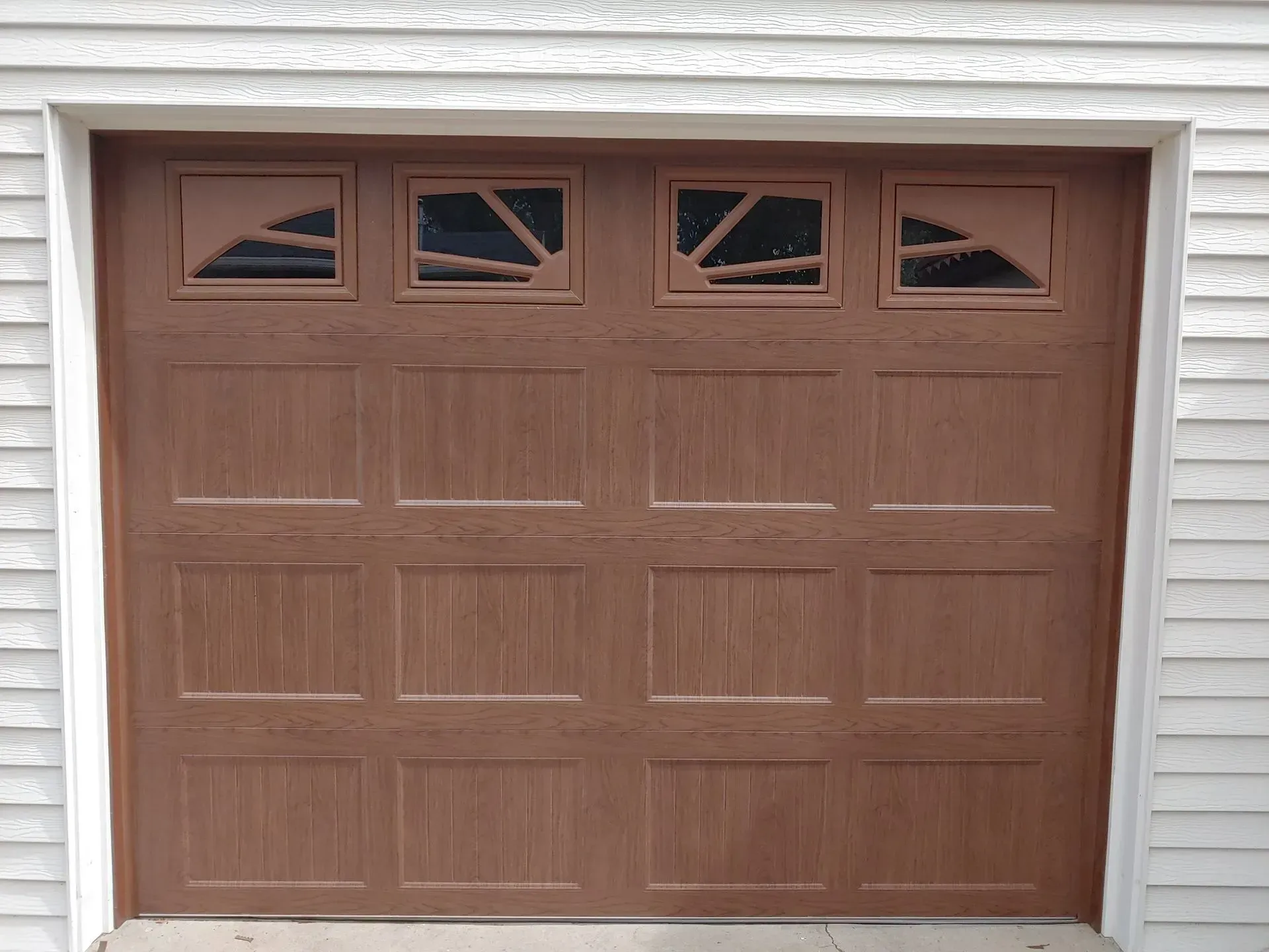 Brown garage door with four windows at the top, set in a white frame on a white siding wall.