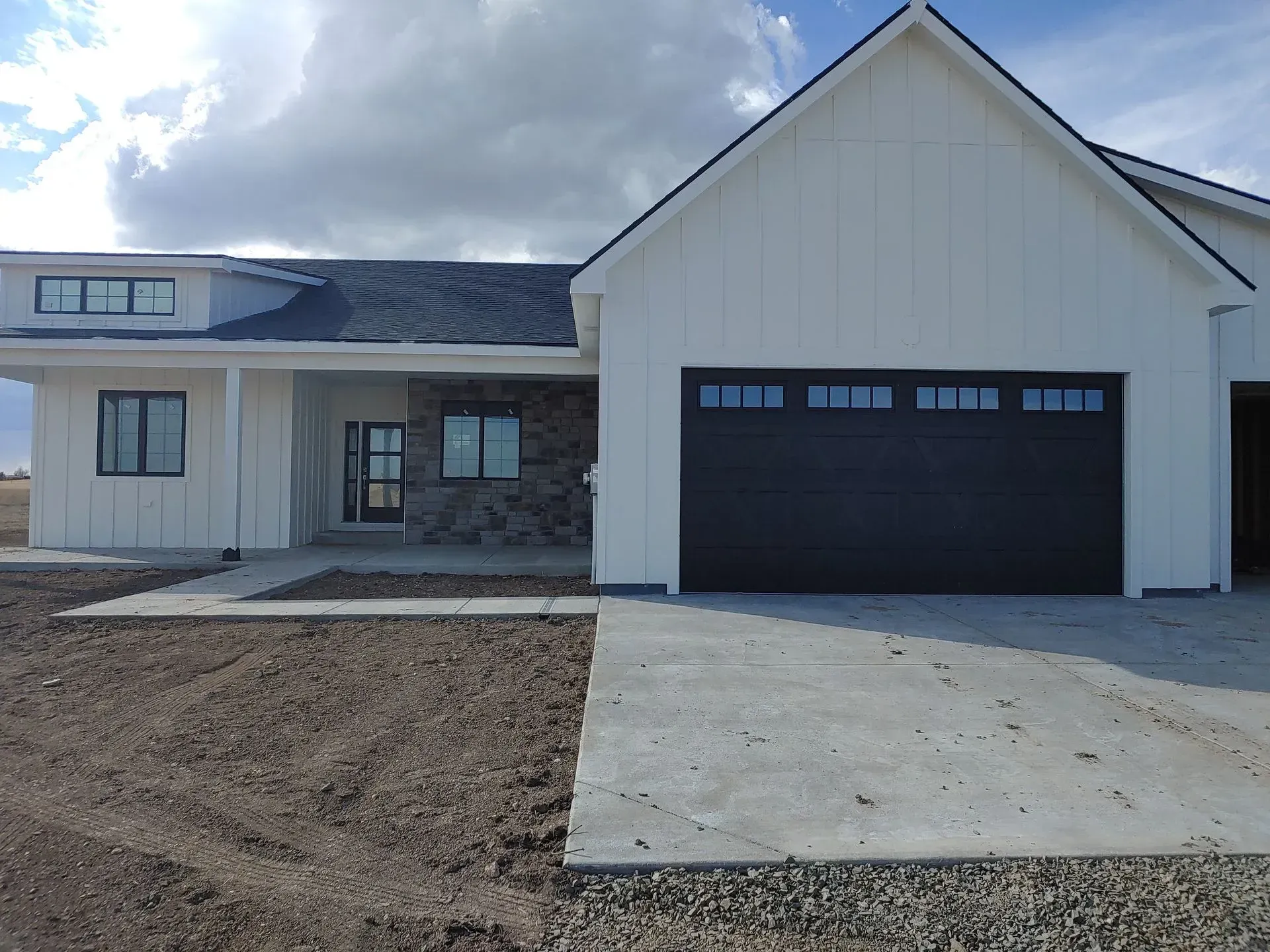 Modern white house with black garage door, stone accents, and concrete driveway on a cloudy day.
