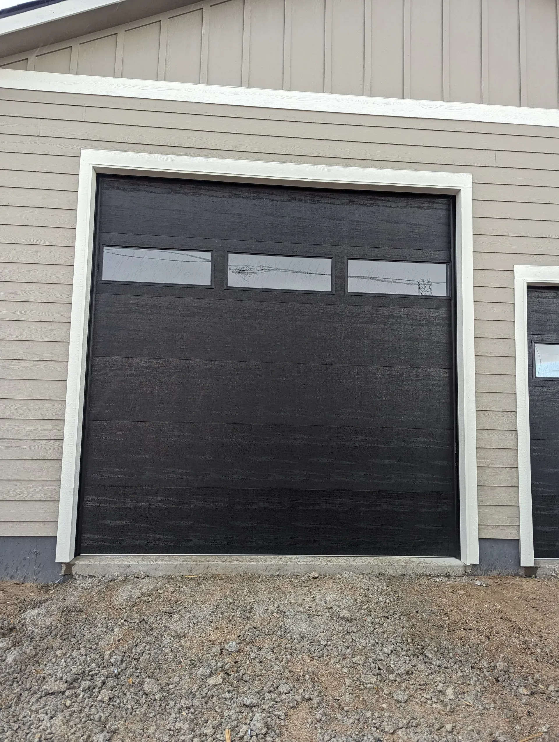Black garage door with three small windows and white trim on a building with light siding.