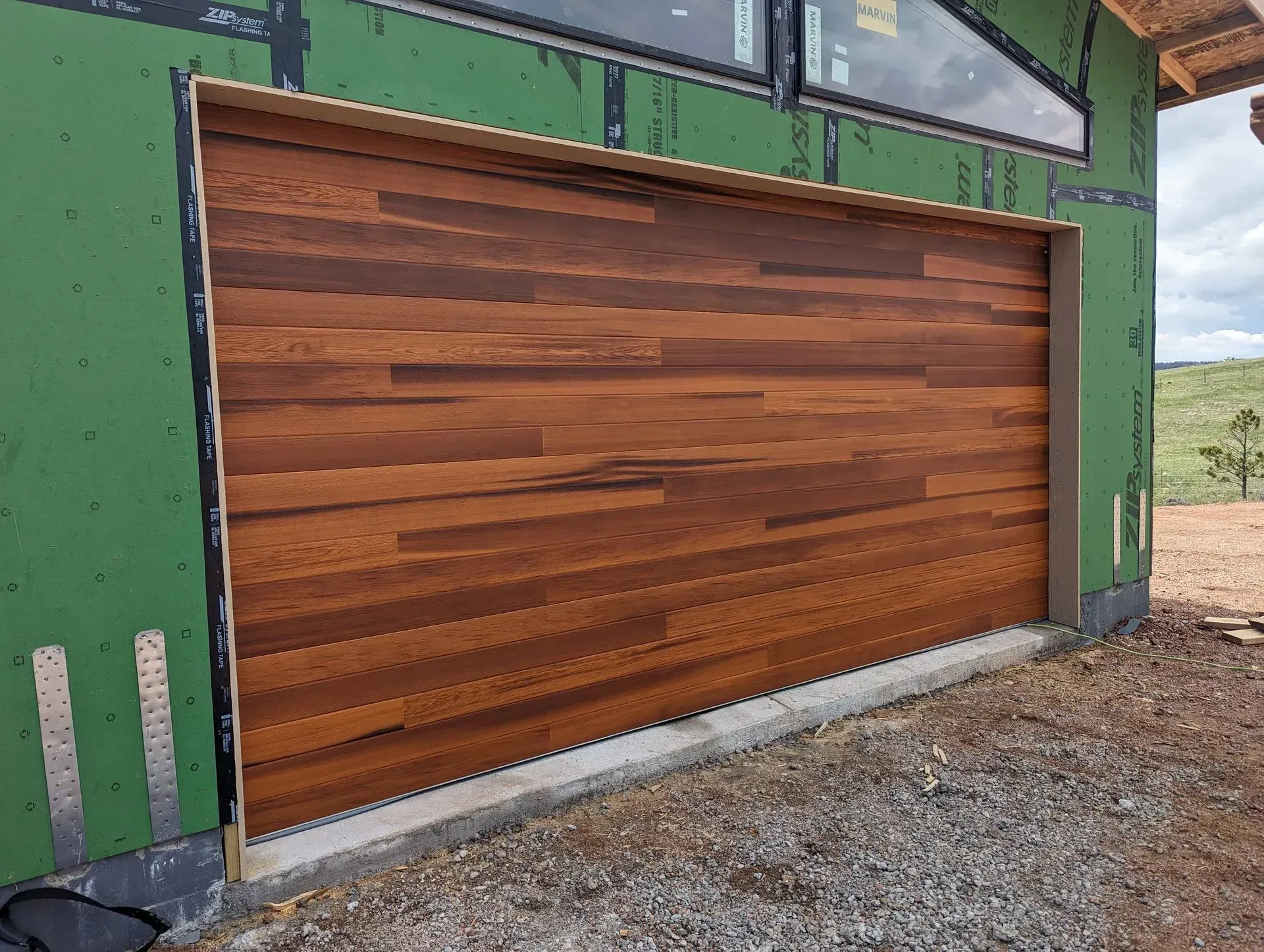 Wooden garage door on a partially built house, framed with light-colored trim. Green siding and a window are above.