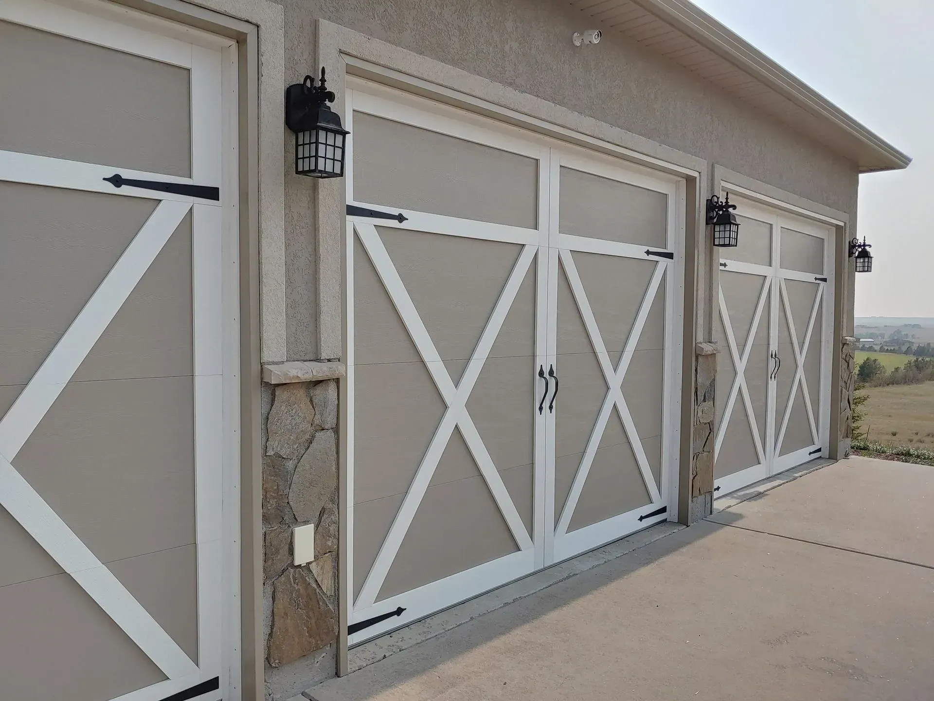 Tan and white garage doors with decorative X design, black hardware, and outdoor lighting.