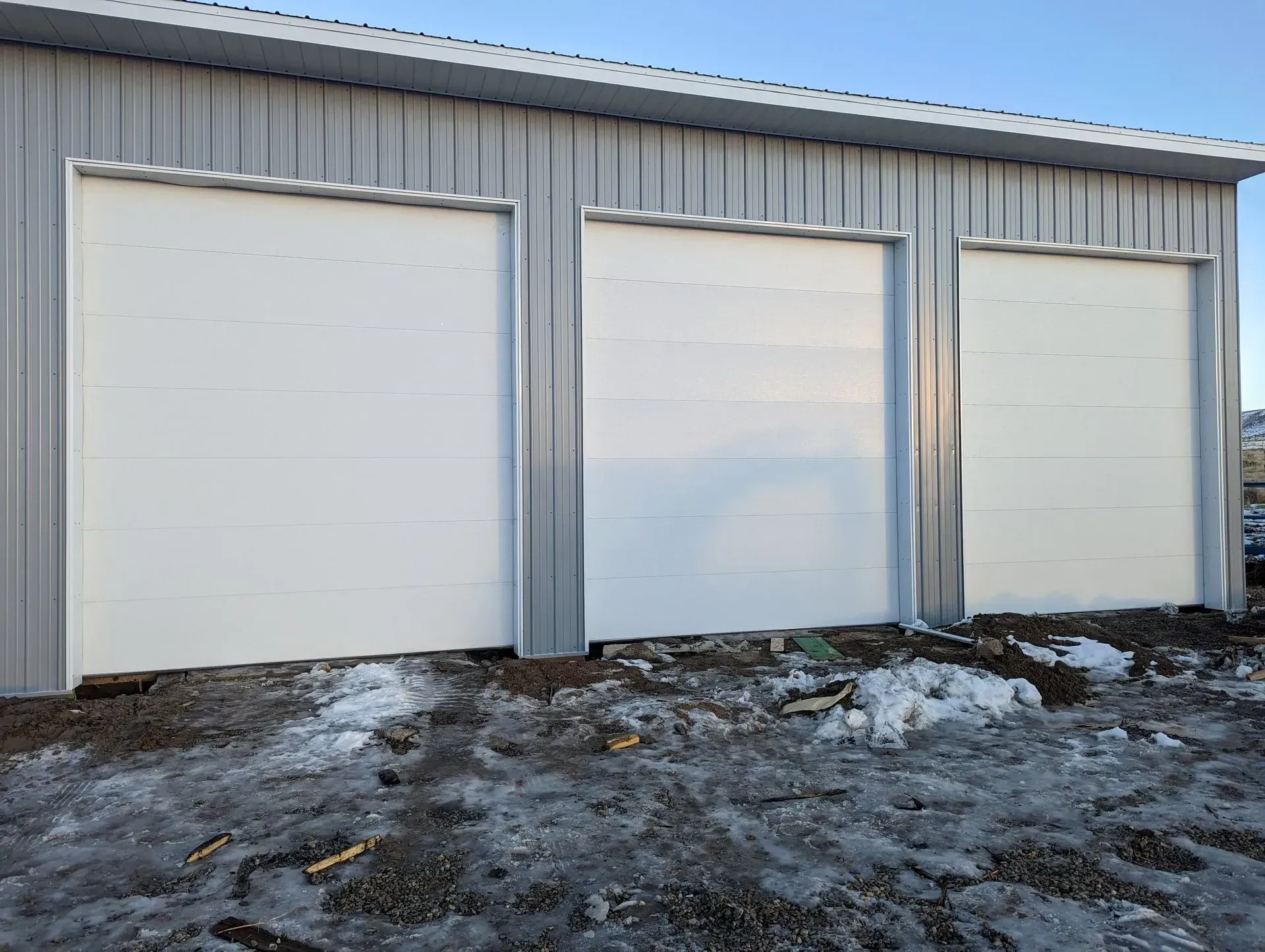 Three white garage doors on a gray metal building exterior. Ground has snow and debris.