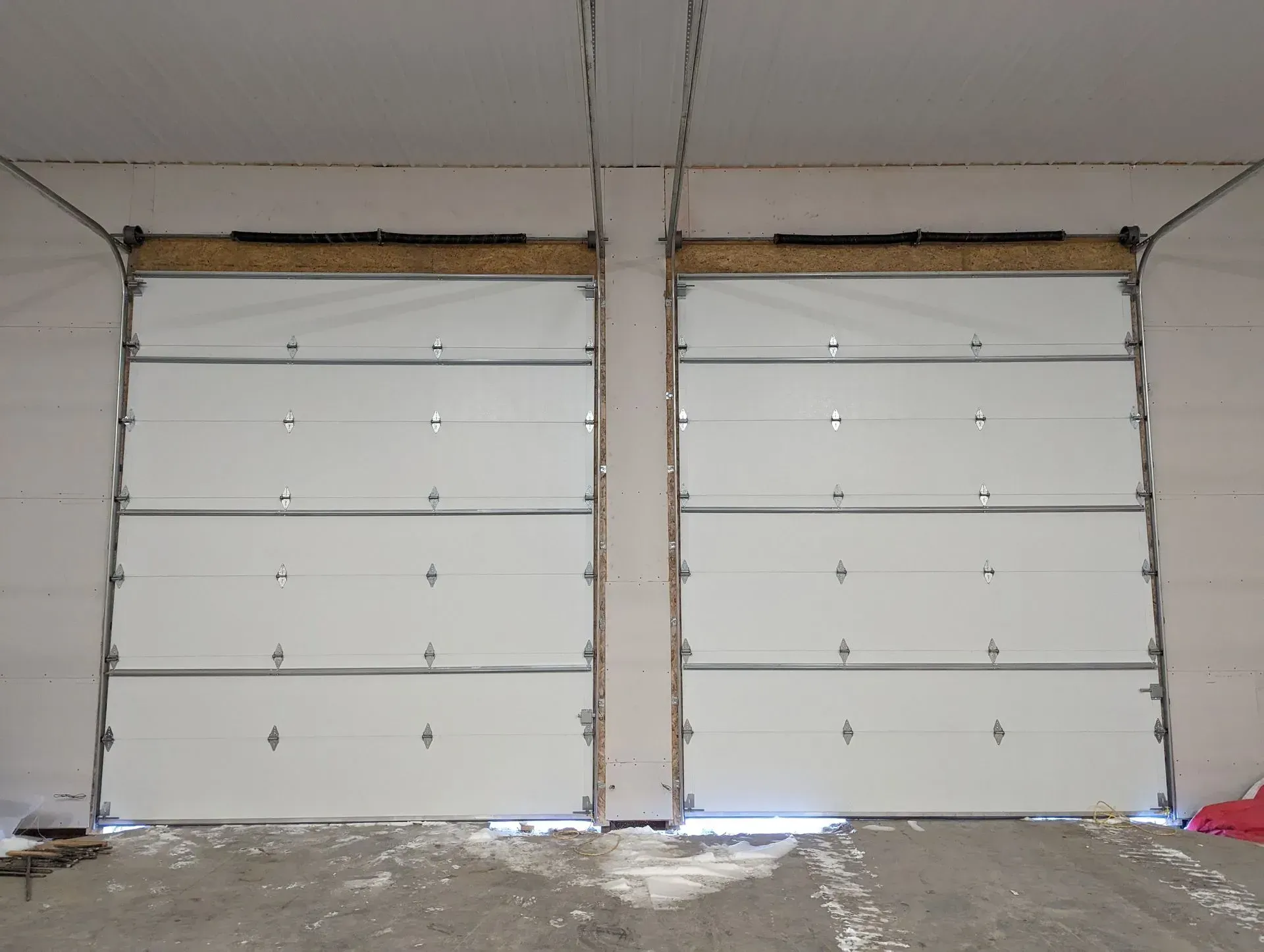 Two white overhead garage doors in a concrete room, closed, with metal trim and light above.