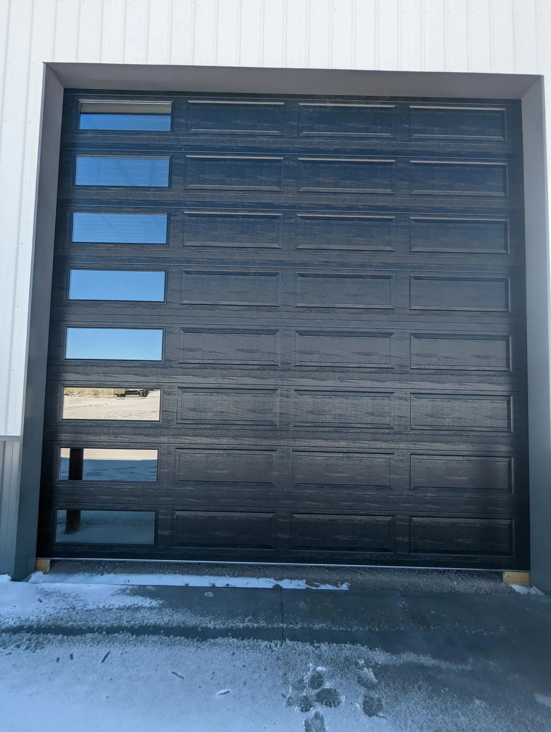 Dark garage door with horizontal texture and rectangular windows. Snow on the ground.