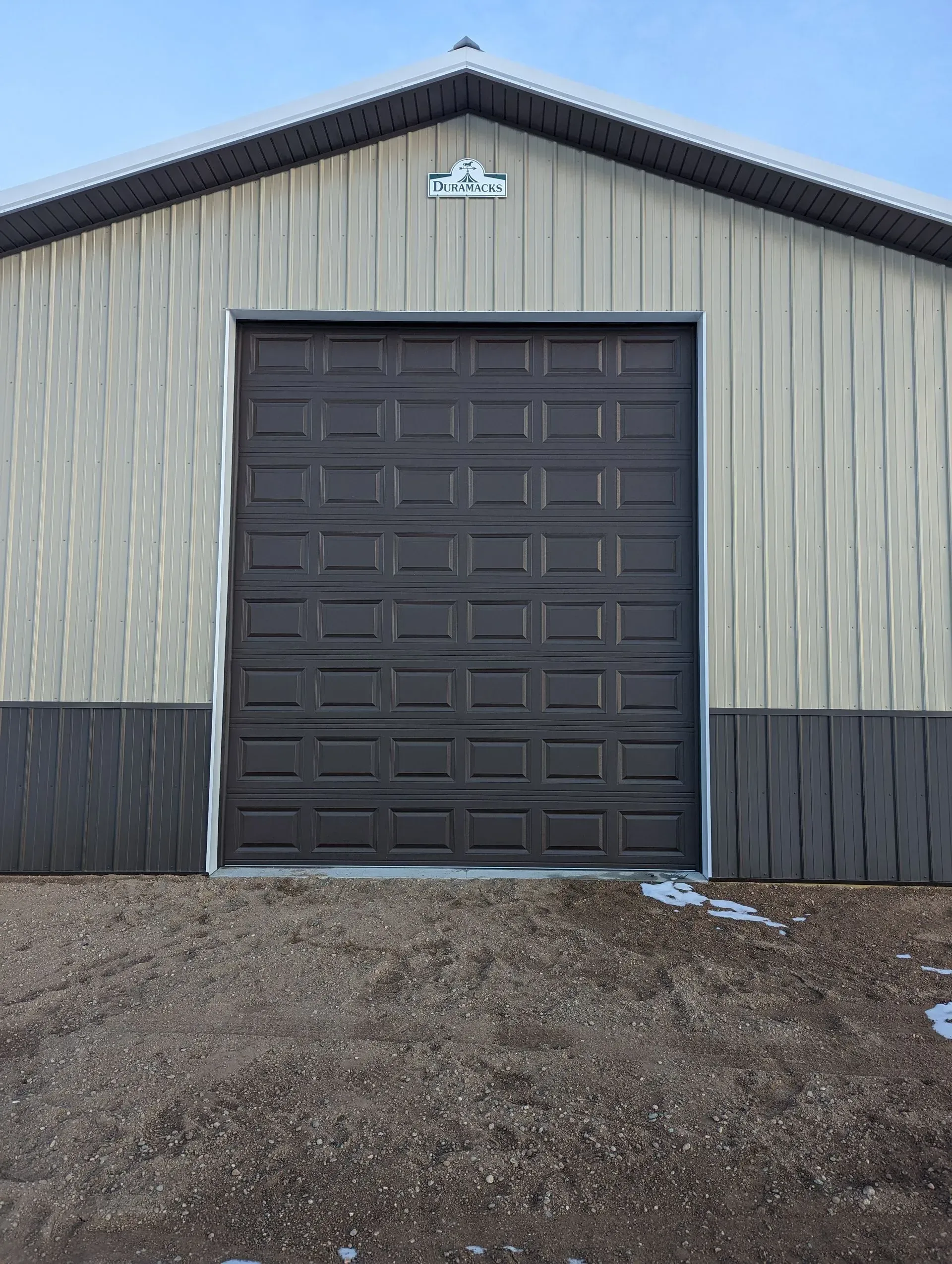 Brown garage door on a beige and brown metal building with a gravel driveway.