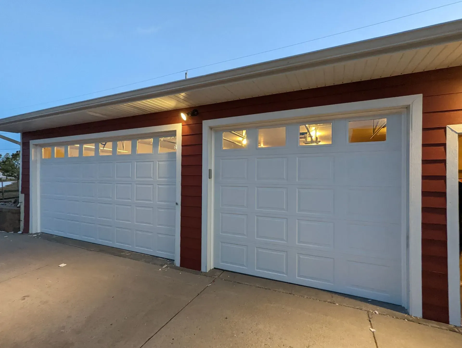 Two white garage doors on a red building, lit up at dusk.