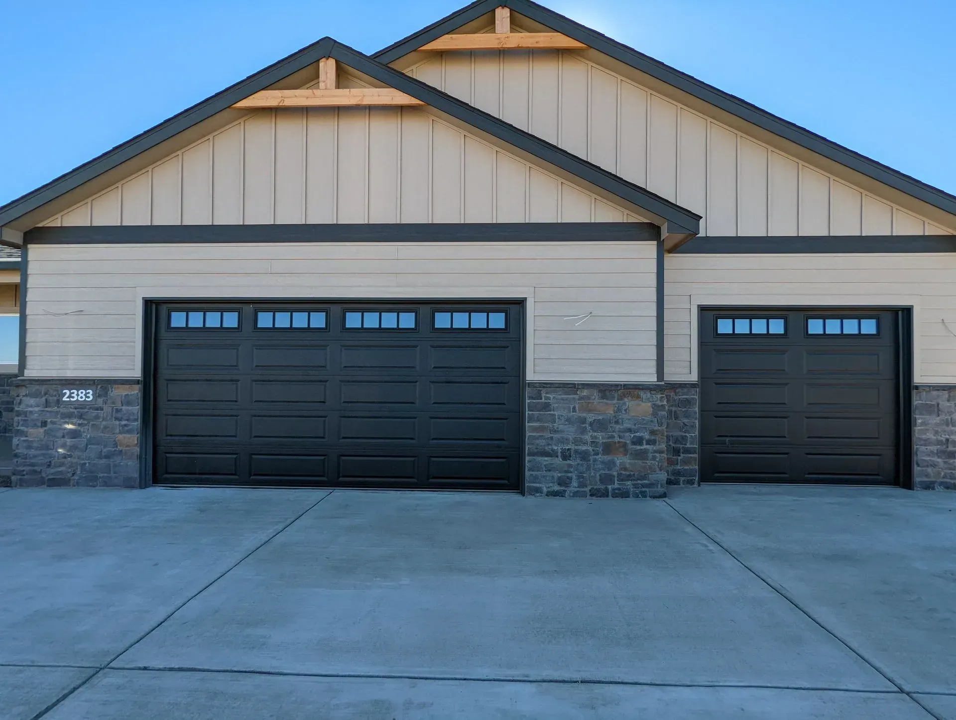 Two-car garage with black doors, stone accents, and tan siding under a clear blue sky.