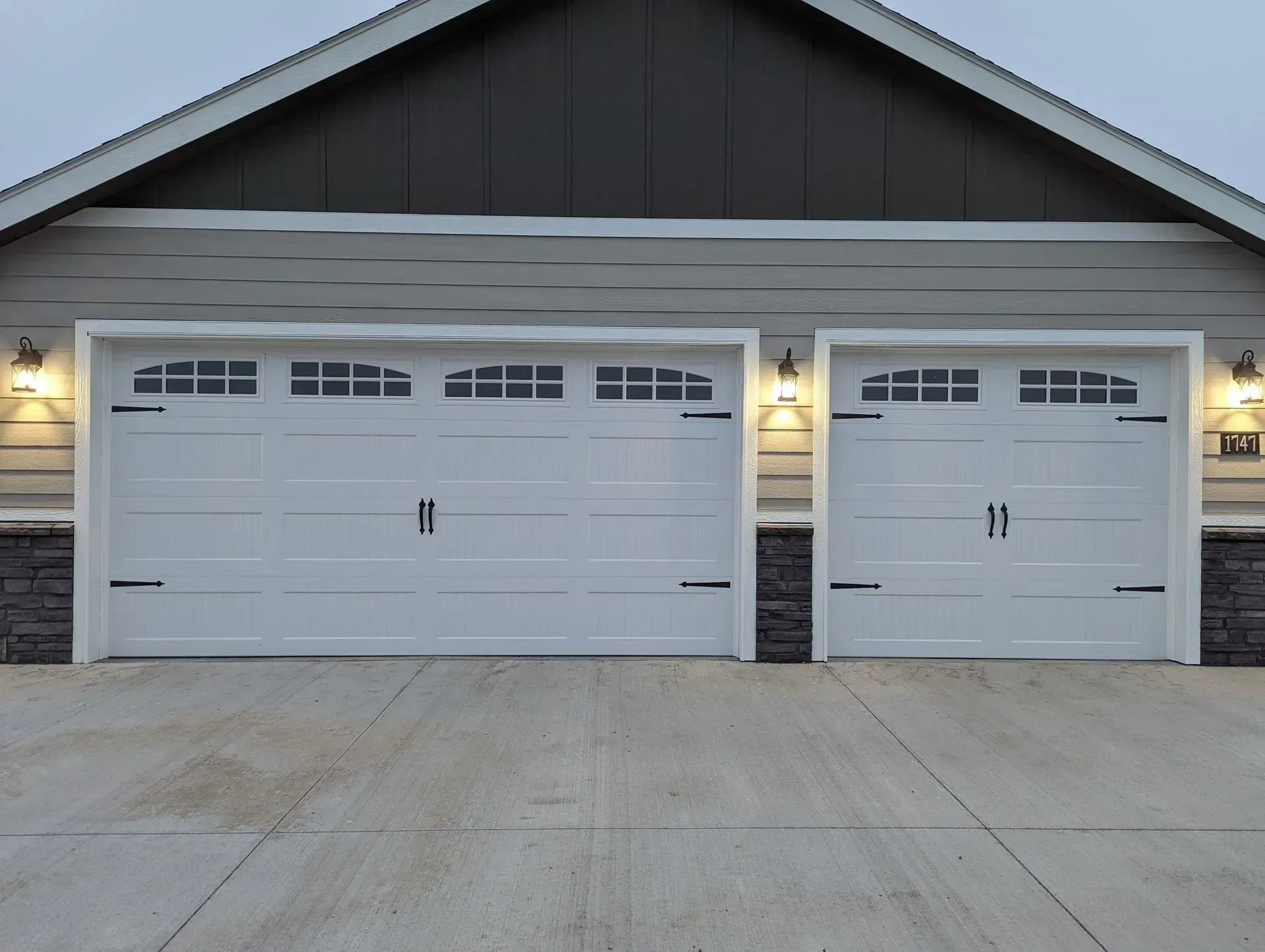 Two white garage doors with black hardware, flanked by stone and siding, with exterior lights.