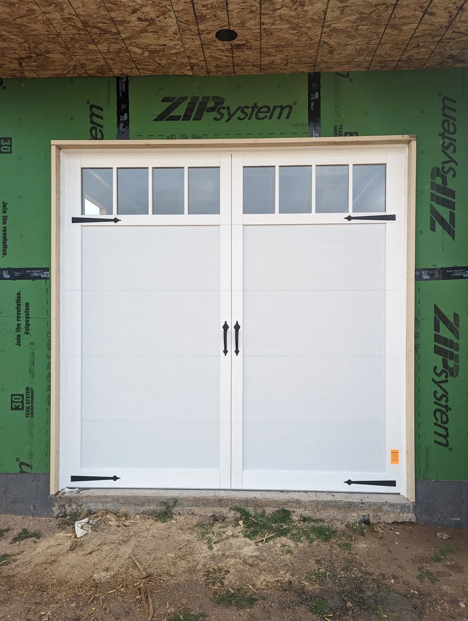 White double garage doors with small rectangular windows, black hardware, and green siding.