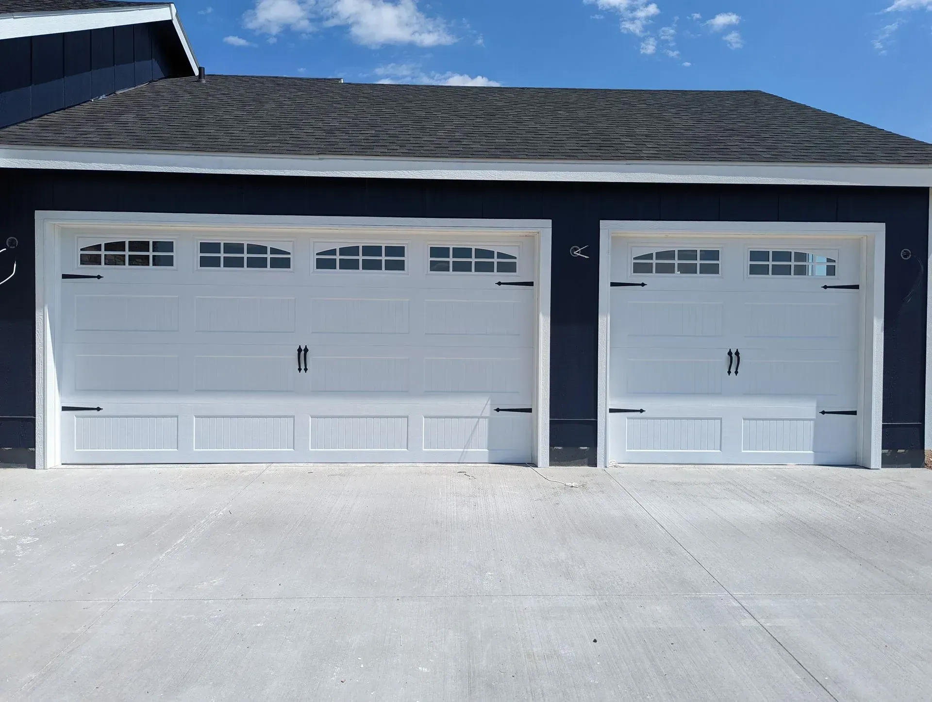 Two white garage doors on a dark blue building, with black hardware, on a concrete driveway.