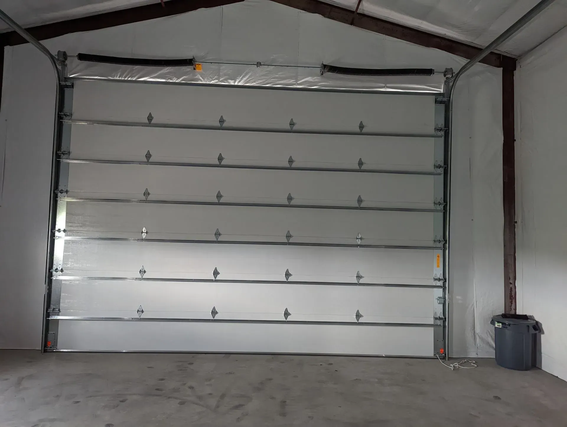 White, sectional garage door in a metal building. Gray trash can to the right.