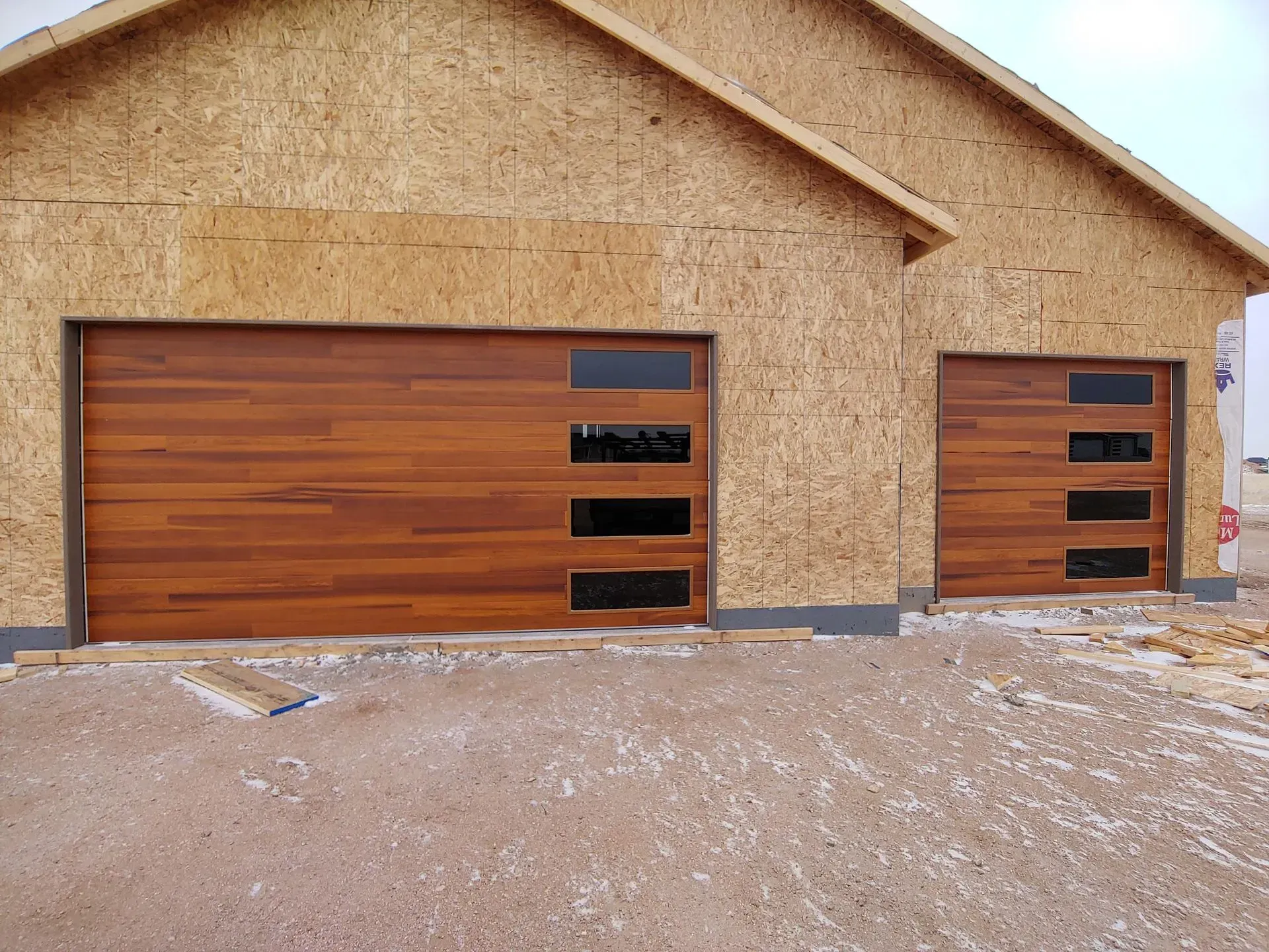 Two wooden garage doors with black windows on a house under construction.