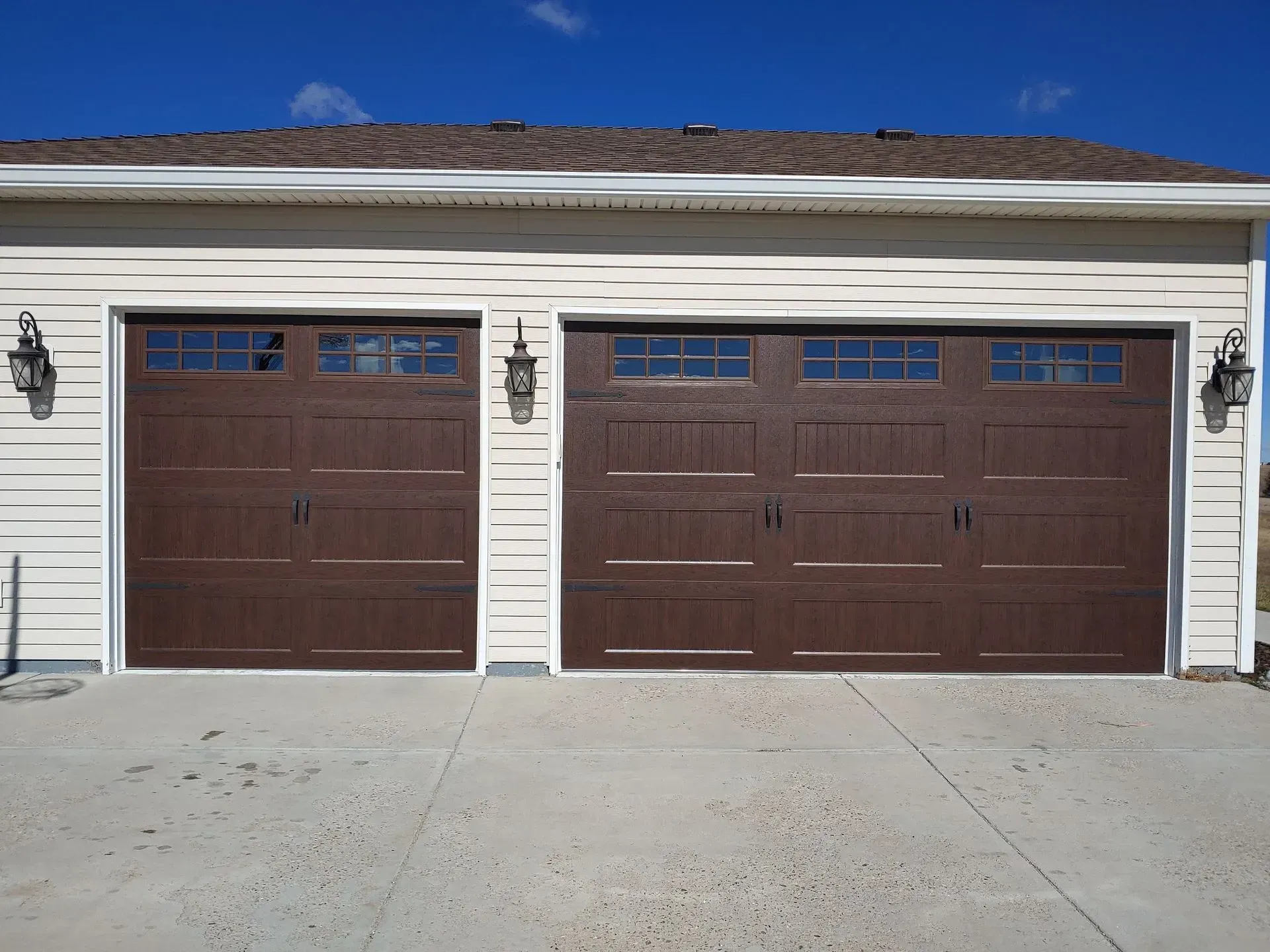 Brown double garage doors with windows, tan siding, and a concrete driveway under a blue sky.
