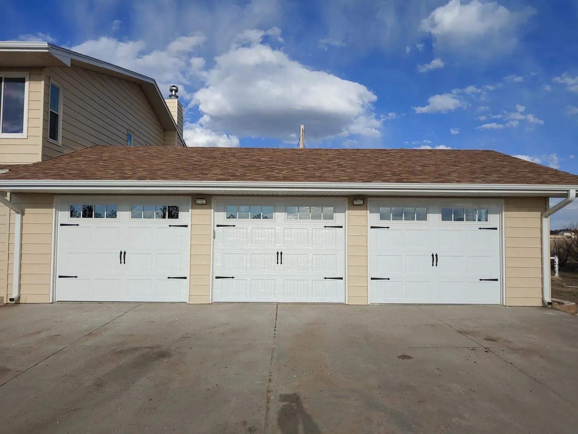 Three-car garage with white doors, tan siding, and a brown roof against a blue sky with fluffy clouds.