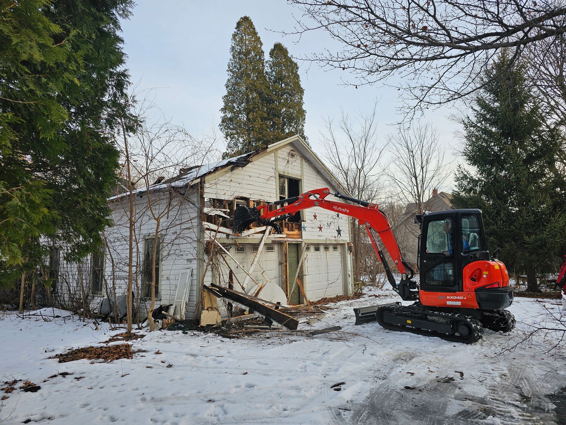 Orange excavator demolishing a small white building in a snowy yard