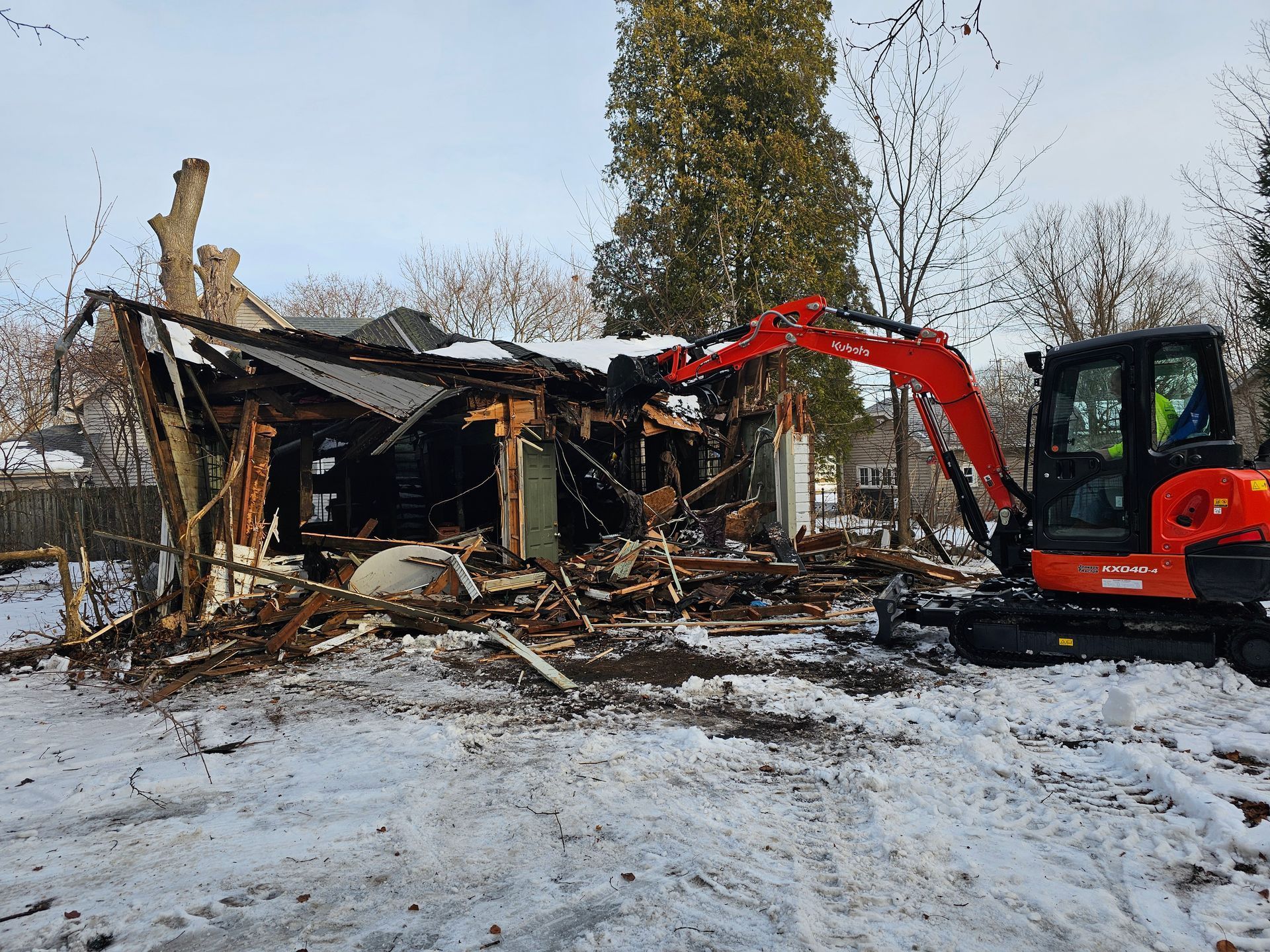 Collapsed house ruins with a red excavator clearing debris in a snowy yard