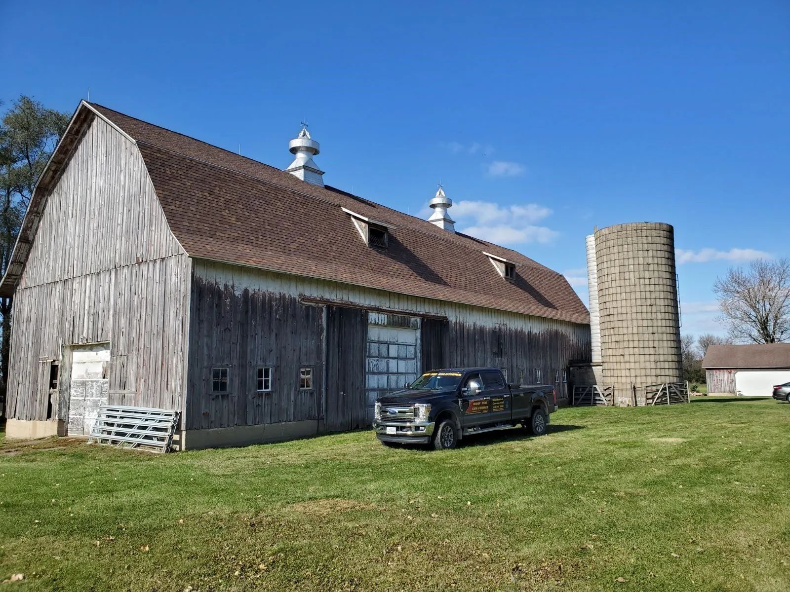 Rustic barn with a parked black truck and silo on a sunny rural farmyard.