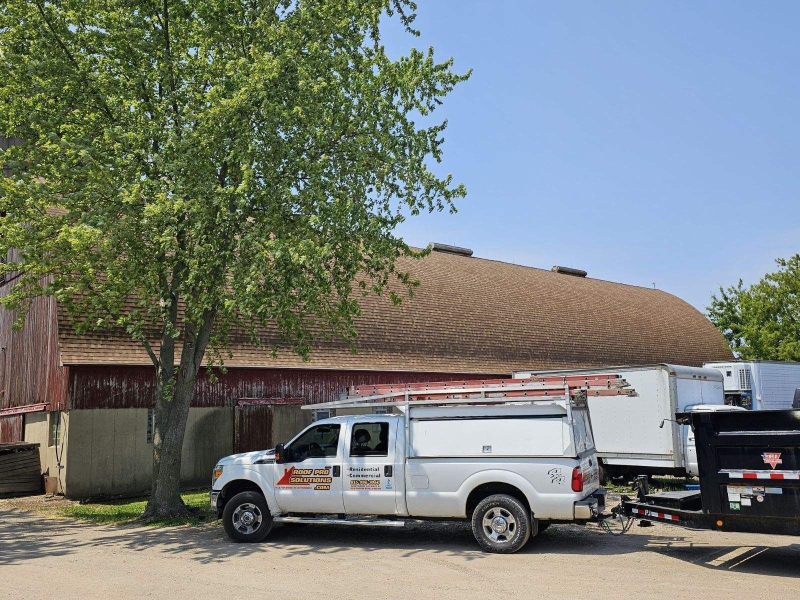 White utility truck parked beside a tan building under a leafy tree on a sunny day