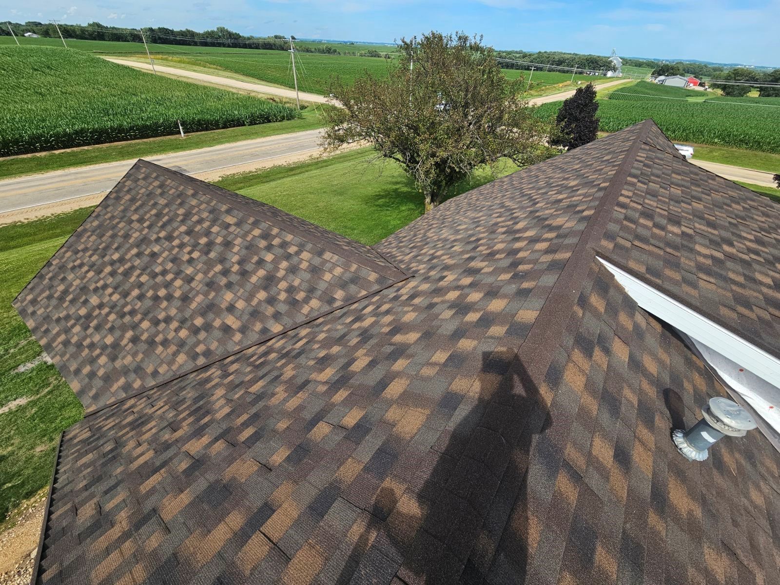 View from a rooftop with brown shingles overlooking fields and a road under a clear sky.
