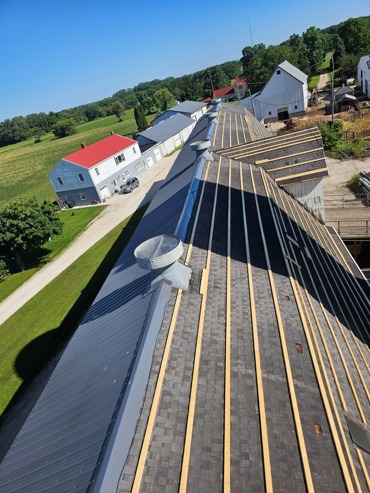 Aerial view of a rooftop under construction with black panels, wooden battens, and nearby farm buildings