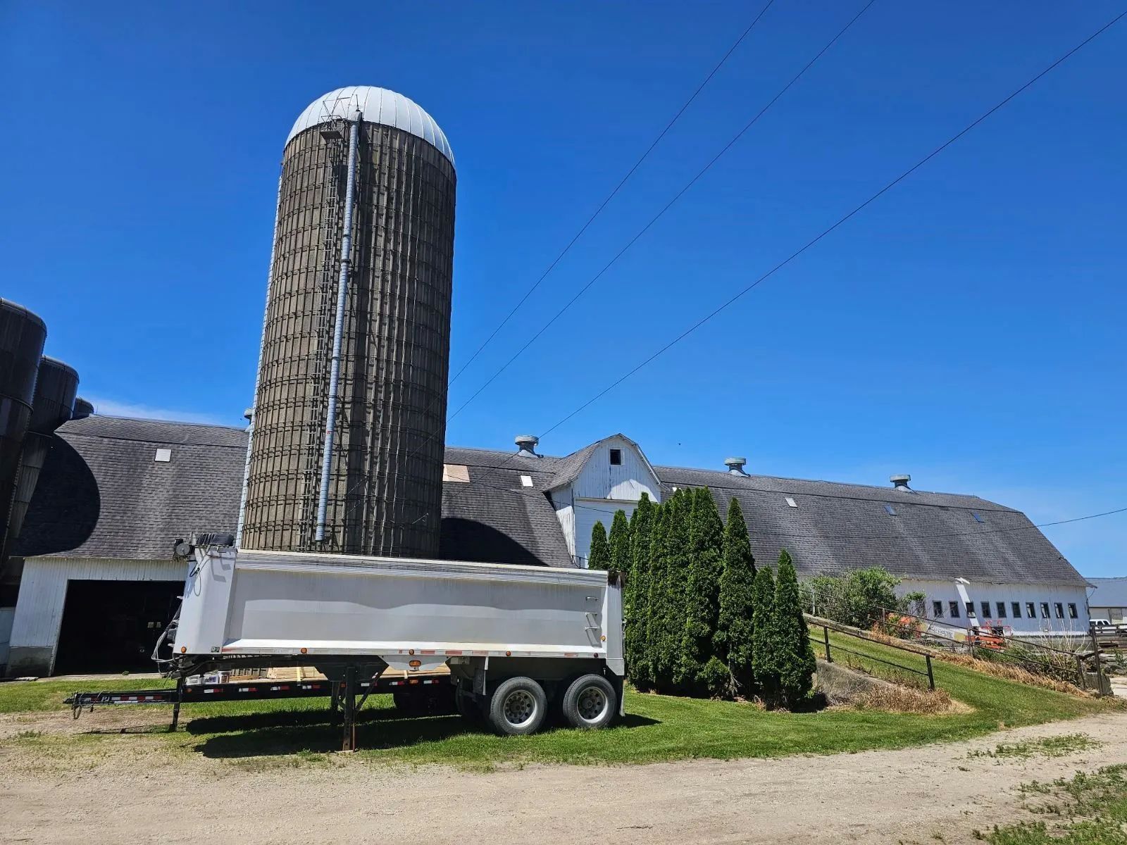 Tall cylindrical silo beside a barn under a clear blue sky, with a white trailer in front.