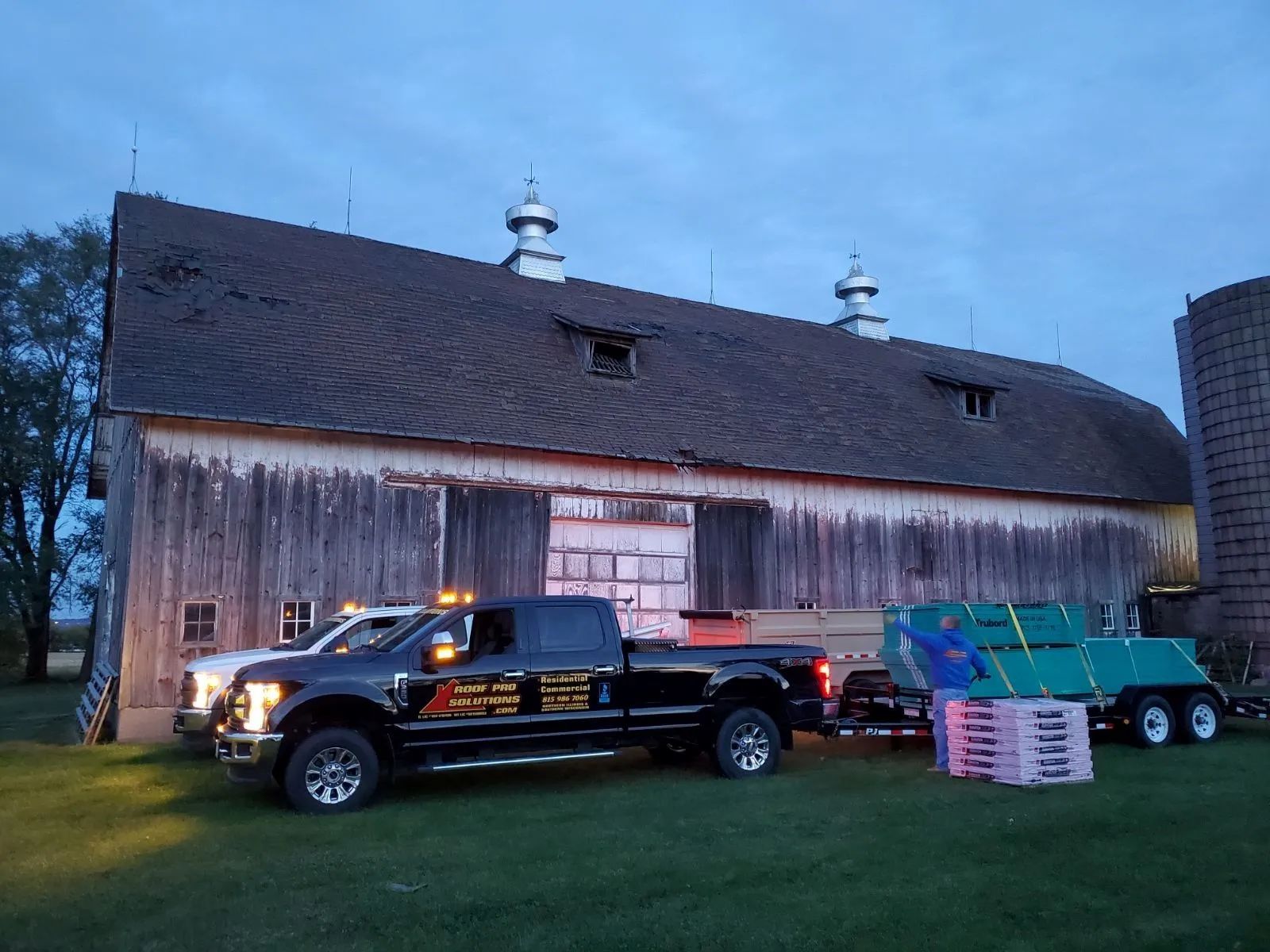 Black pickup truck towing a green trailer beside a weathered barn at dusk