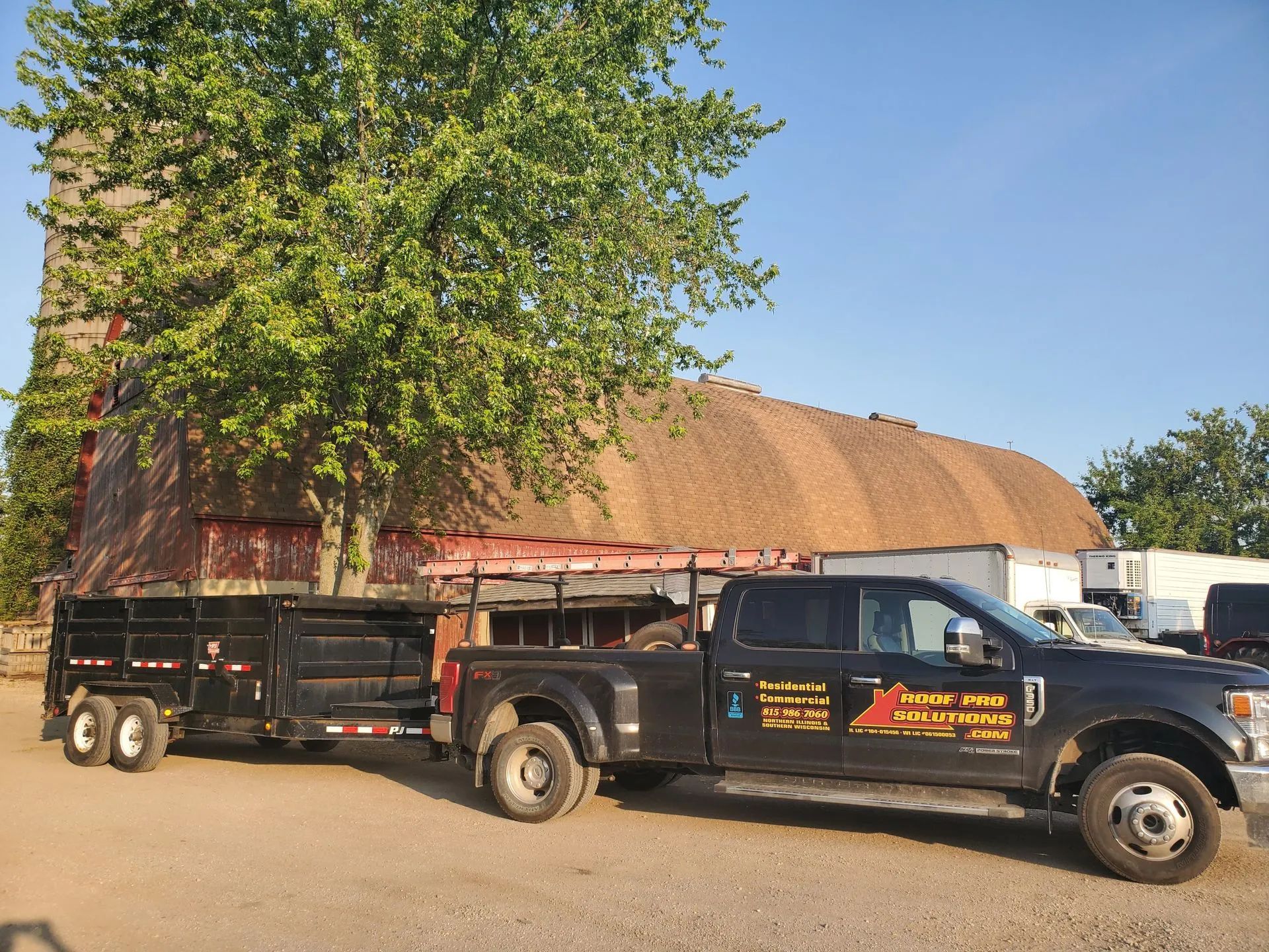 Black pickup truck towing a trailer beside a large tree and rustic building under a clear blue sky