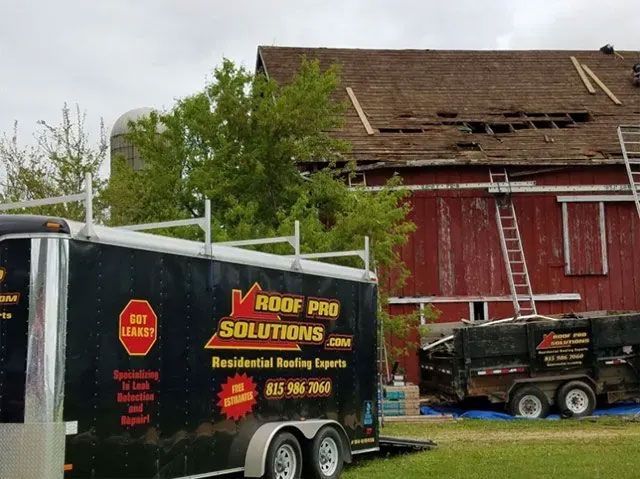 Roofing company trucks parked beside a damaged barn with missing roof tiles and a tree in front