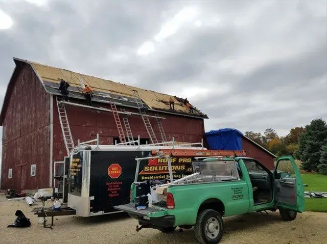 Workers repairing a barn roof beside a green pickup and ladder under a cloudy sky