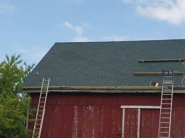 Red barn roof under repair with two ladders leaning against it and cloudy sky overhead