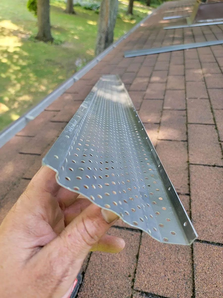 Hand holding a perforated metal flashing strip on a shingled roof with trees in the background