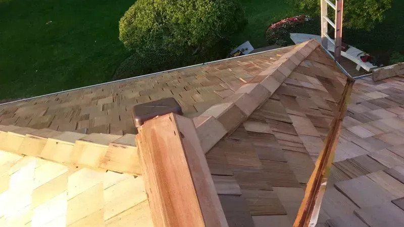 Roofing under construction with tan shingles and a wooden ridge against a green yard below