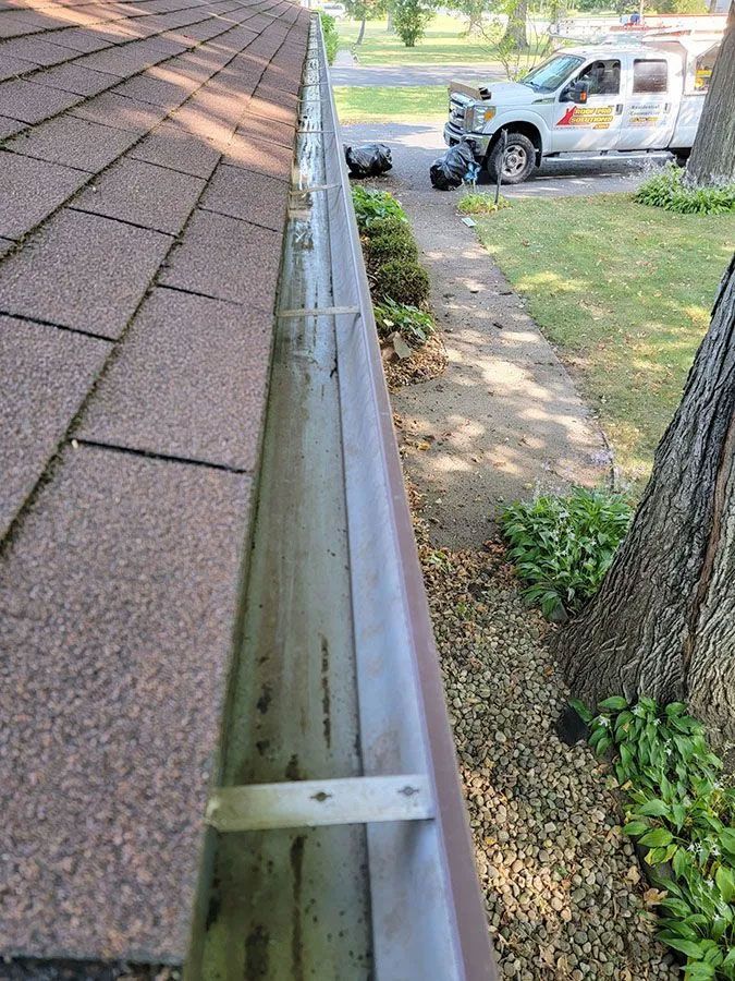 Gutter along a shingled roof beside a tree and lawn, with a white service truck in the background