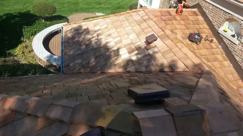 Roof under construction with tan shingles, tools, and sunlight casting shadows.