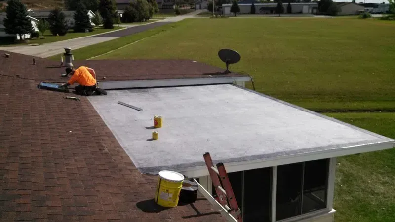 Worker in orange on a roof painting a flat white extension next to a shingled roof.