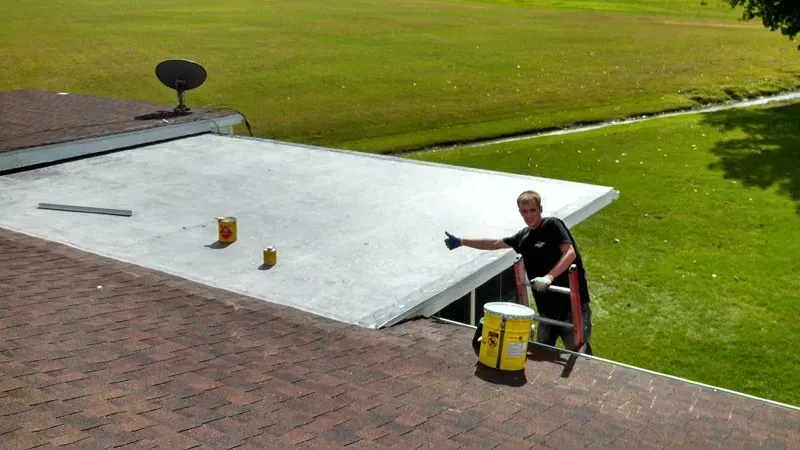 Worker on a flat roof applying white coating beside a green lawn.