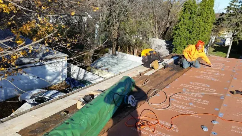 Workers installing roofing underlayment on a sloped roof beside snowy trees.