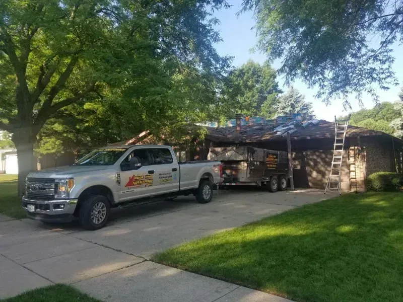 White work pickup truck parked beside a house under trees, with a trailer and yard equipment in the driveway