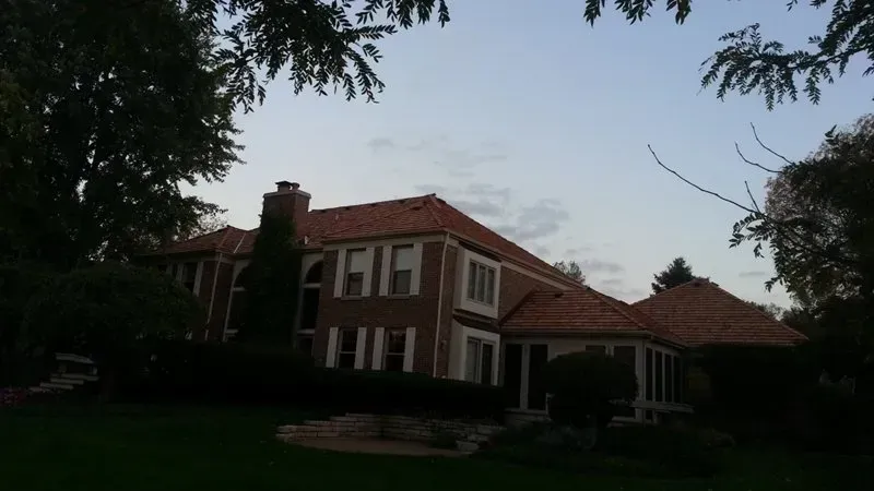 Large brick house with red roof and white trim at dusk, framed by dark trees