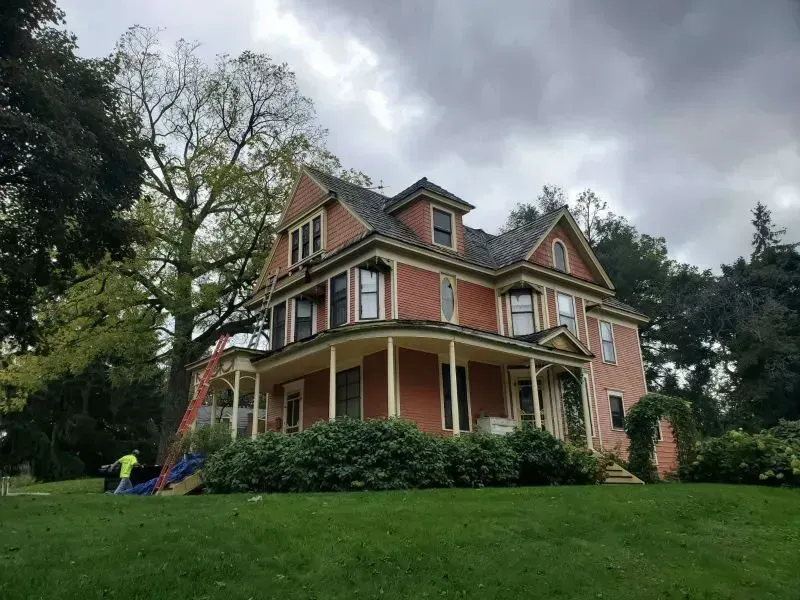 Victorian house with wraparound porch on a grassy hill under a cloudy sky